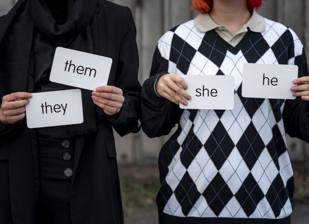 The torso's of two people are seen close up and holding signs naming their pronouns.