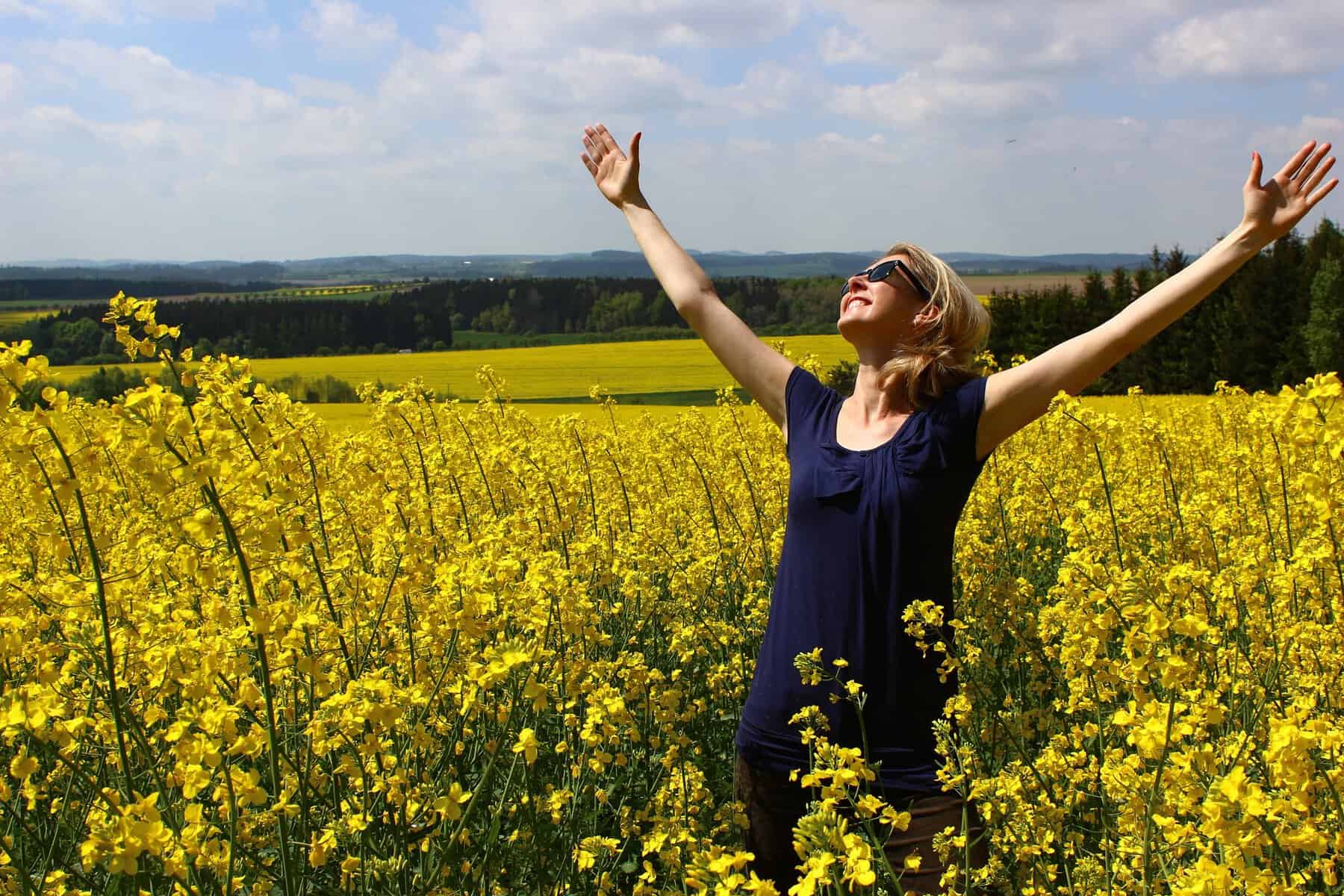 A woman stands in a field of yellow flowers with arms spread wide.