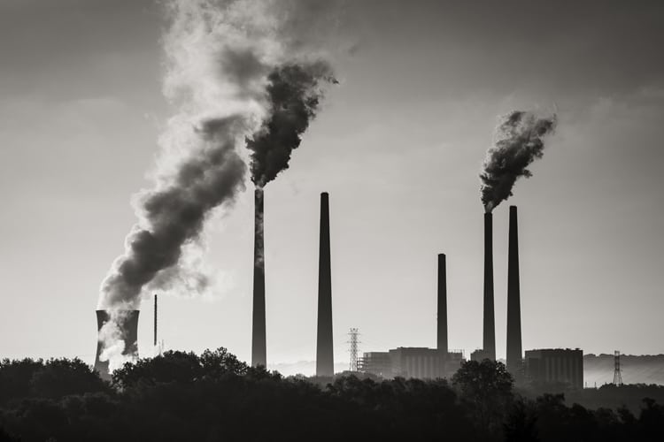 This black and white image shows a group of industrial coal smokestacks releasing smoke into the air.