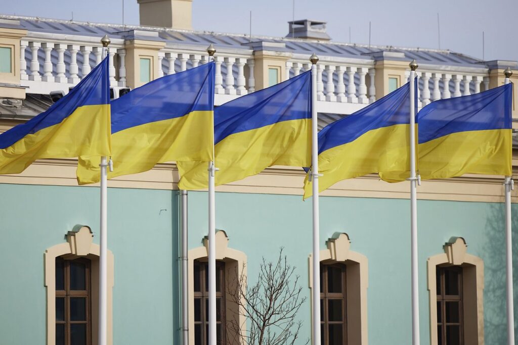 Five flags of Ukraine as seen flying on flagpoles in front of a building.