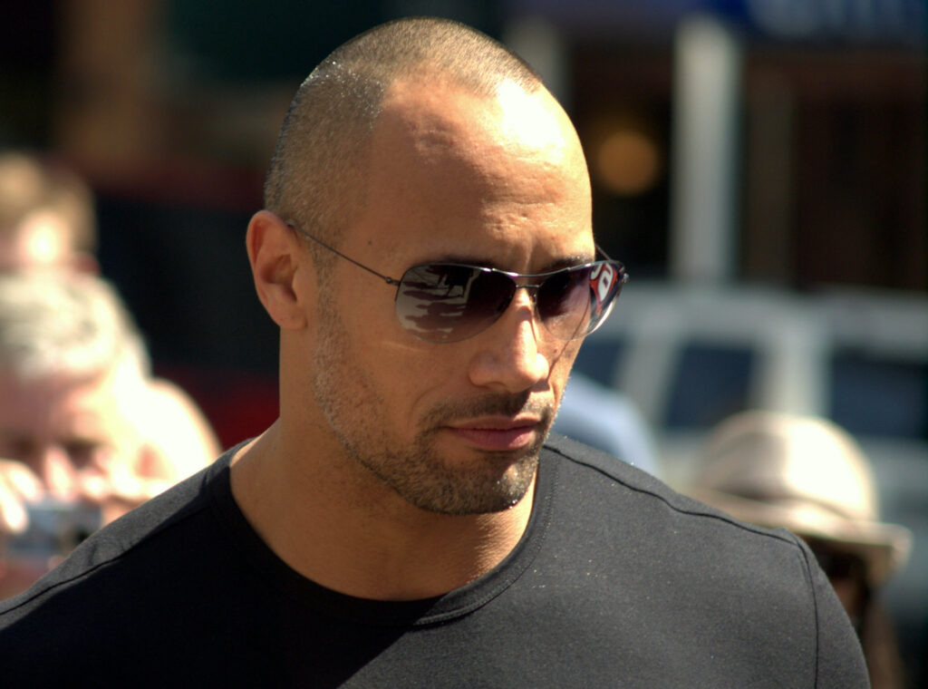 Actor Dwayne Johnson is shown from shoulders up wearing a black T-shirt and sunglasses outdoors.
