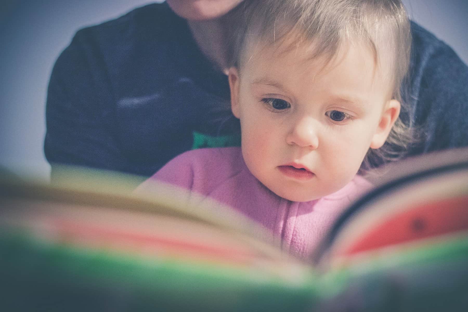 A toddler sits on an adults lap as they look at an open book that is out of focus and between the camera and readers.