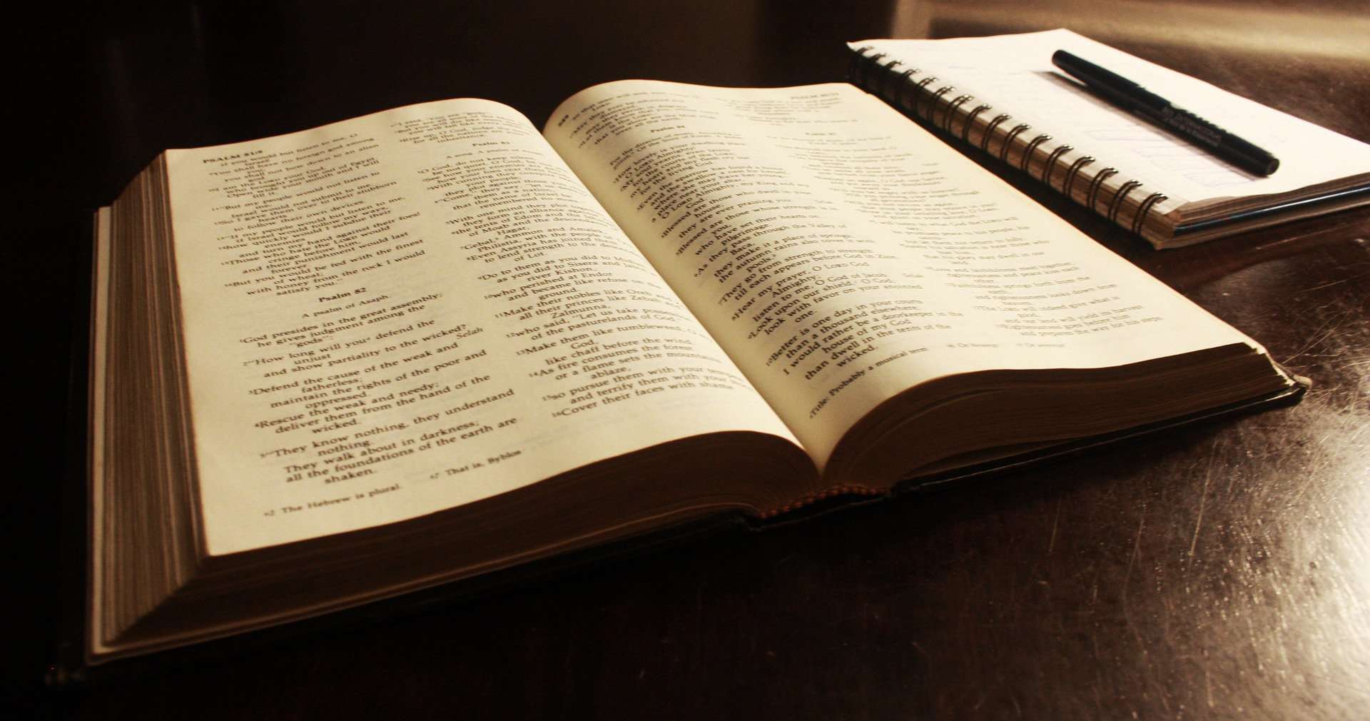 A Bible is shown open faced in soft light with a notepad and with a pen next to it.