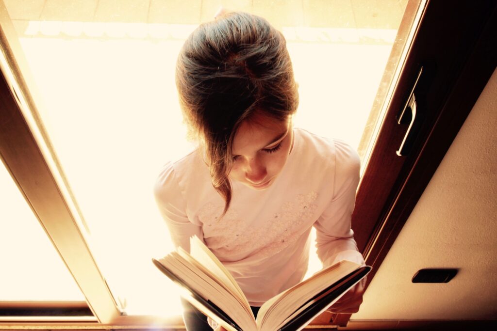 A girl with hair pulled back stands with her back to a glass door and reading a book.