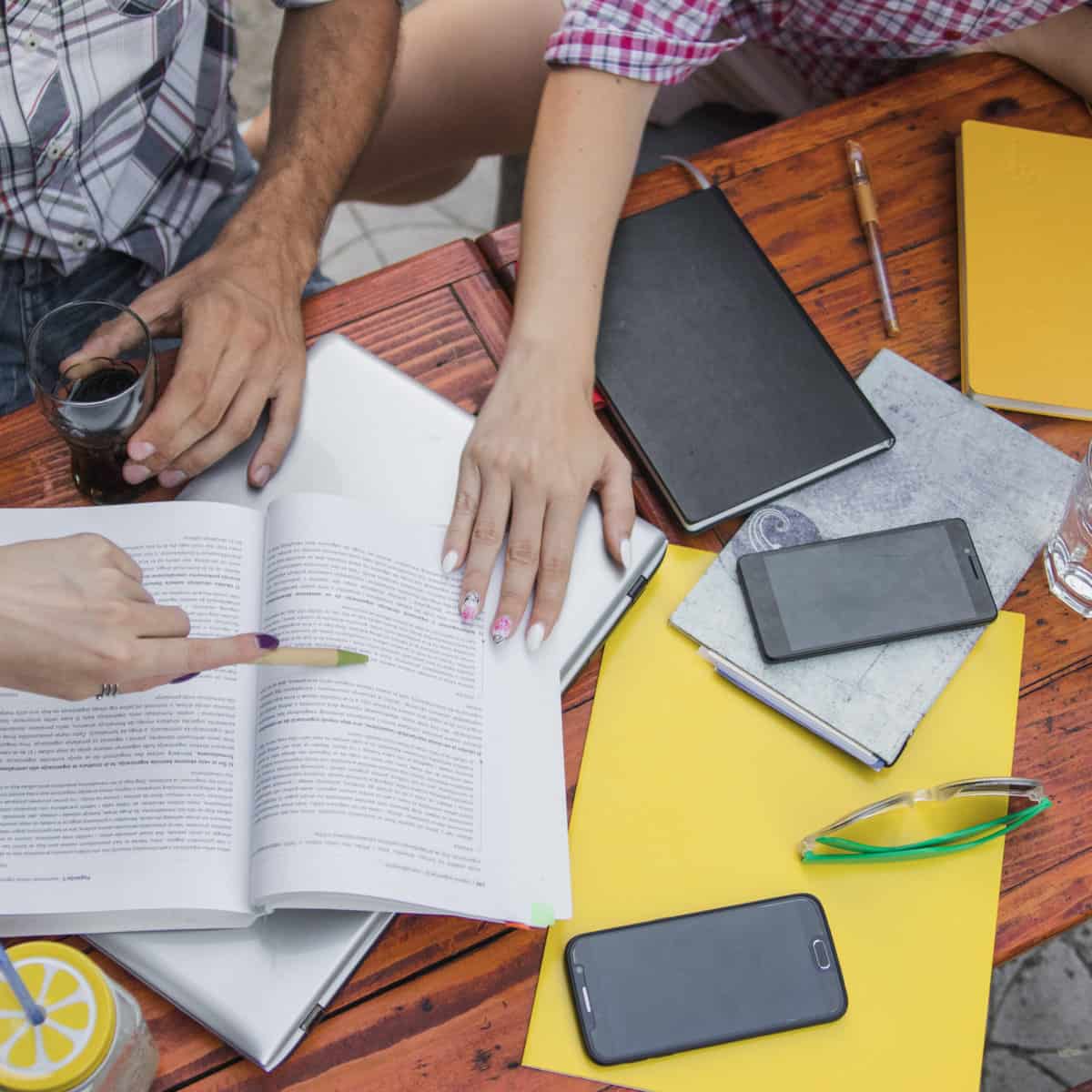 Students sit at a table with an open text book, notepads, cells phones.