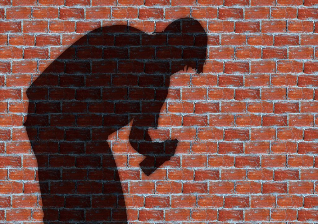The shadow of a man stooping and holding a can of spray paint is seen against a red brick wall.