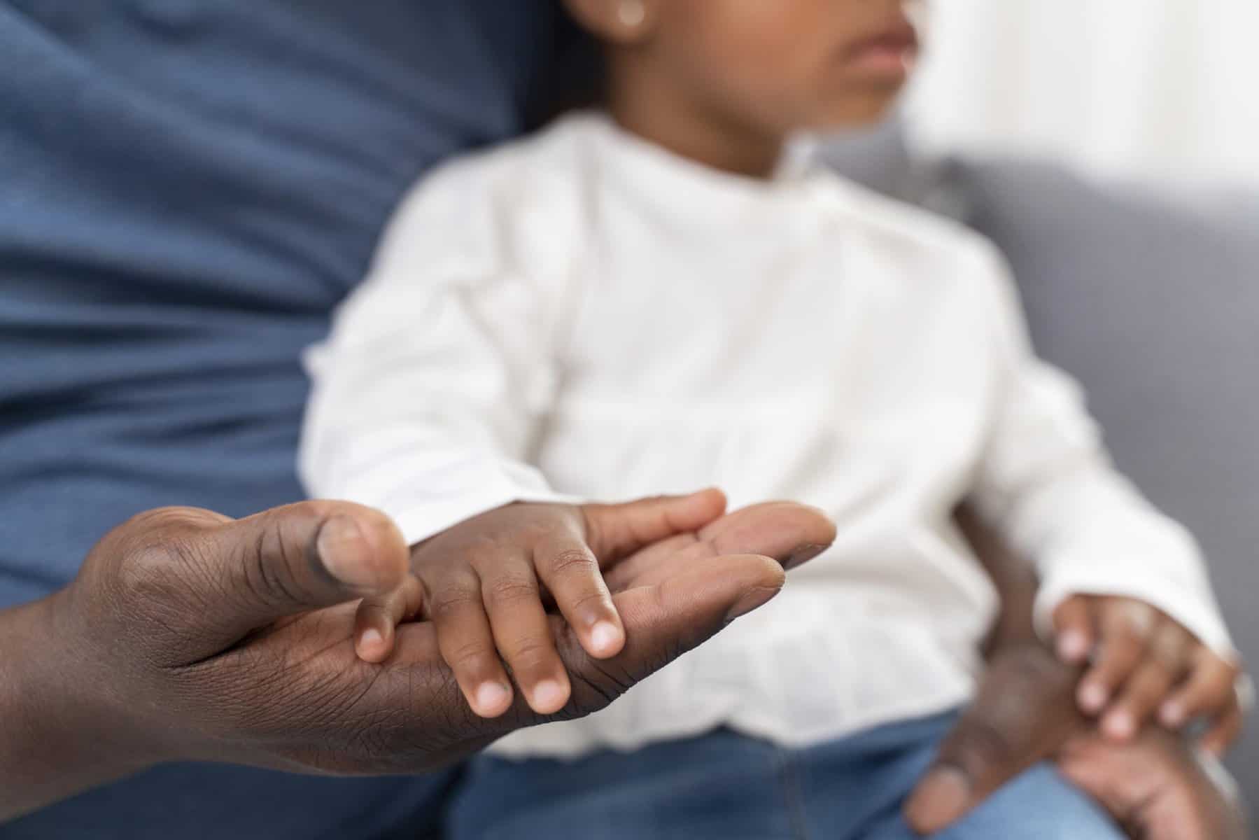 A young Black child sits on the lap of a Black man with hands resting on the man's palm up hands.