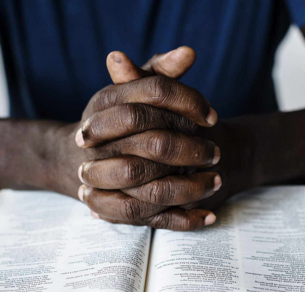 A Black man's hands are shown clasped together and resting on an open Bible.