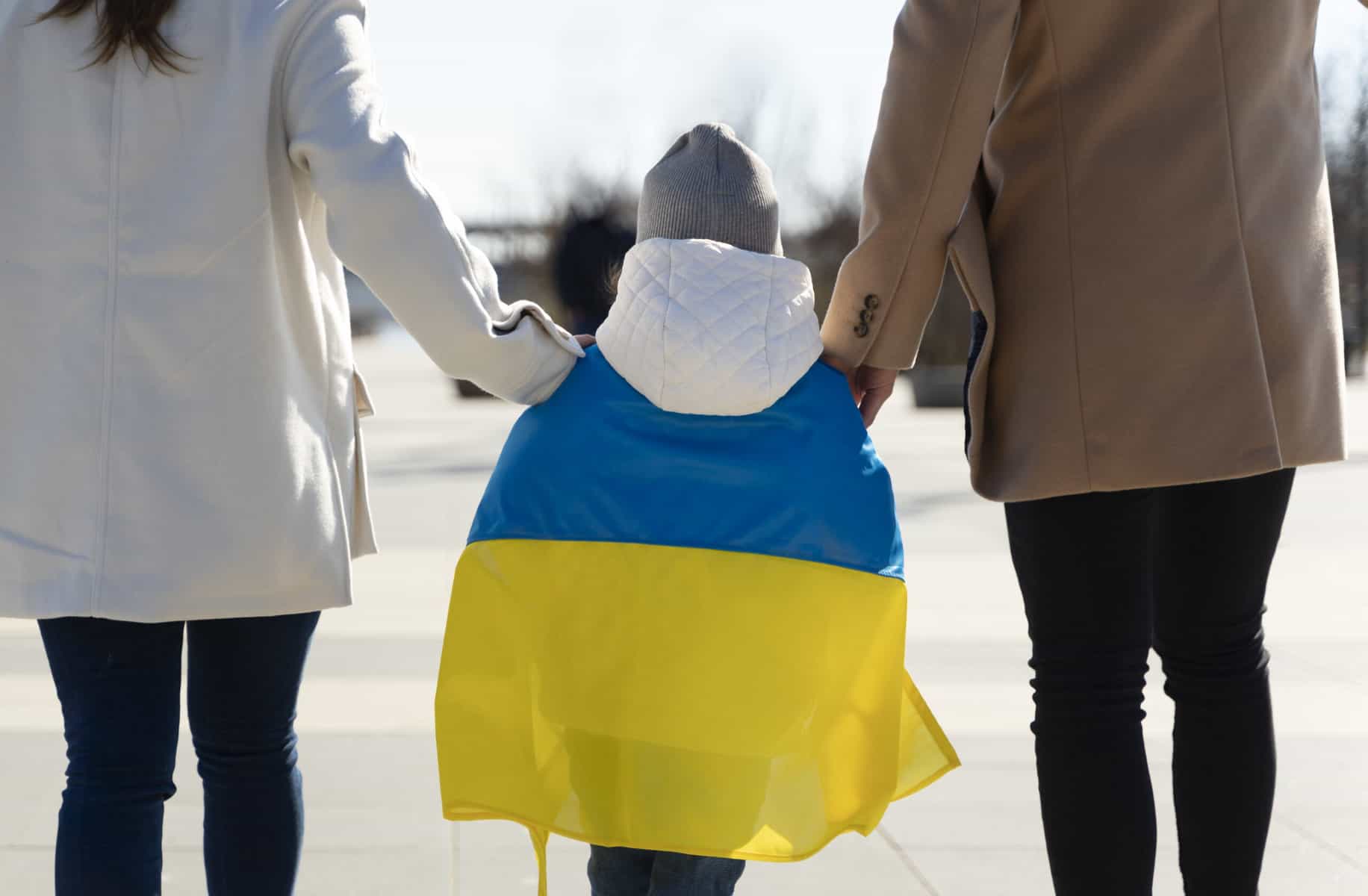 A child walks away from the camera with the flag of Ukraine over their shoulder walks between a man and a woman, holding their hands.