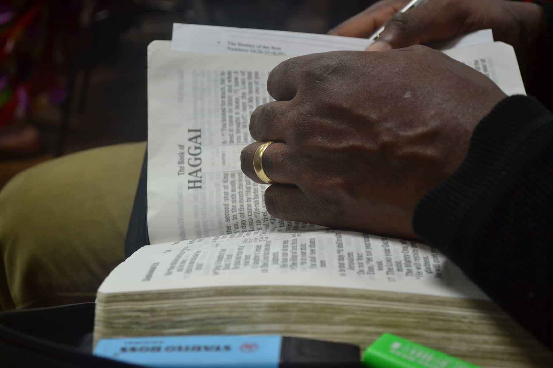 A Black man's hands are seen thumbing through a Bible showing the cover page for the book of Haggai.