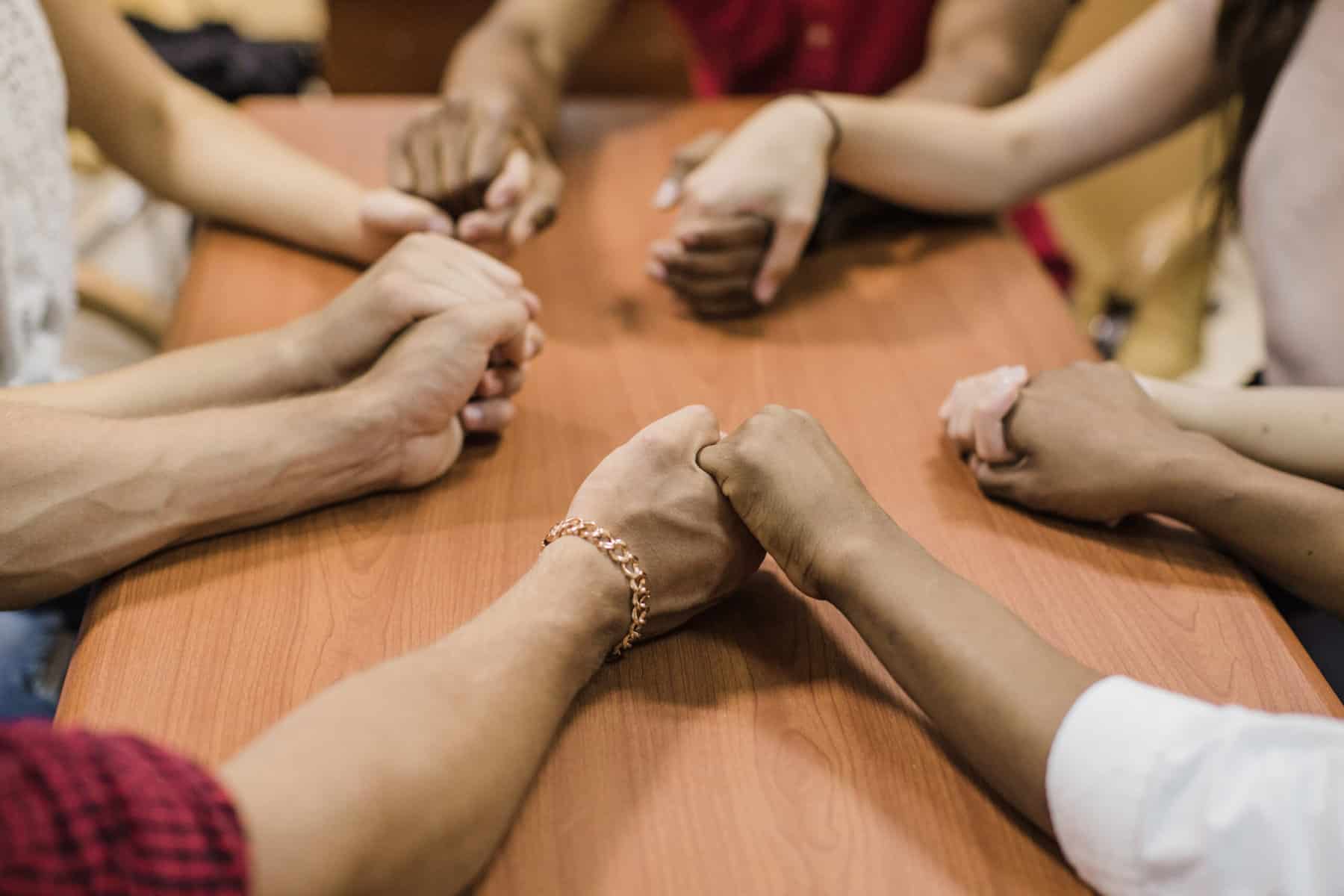 The arms of several people are shown around a small table holding the hands of their neighbors.