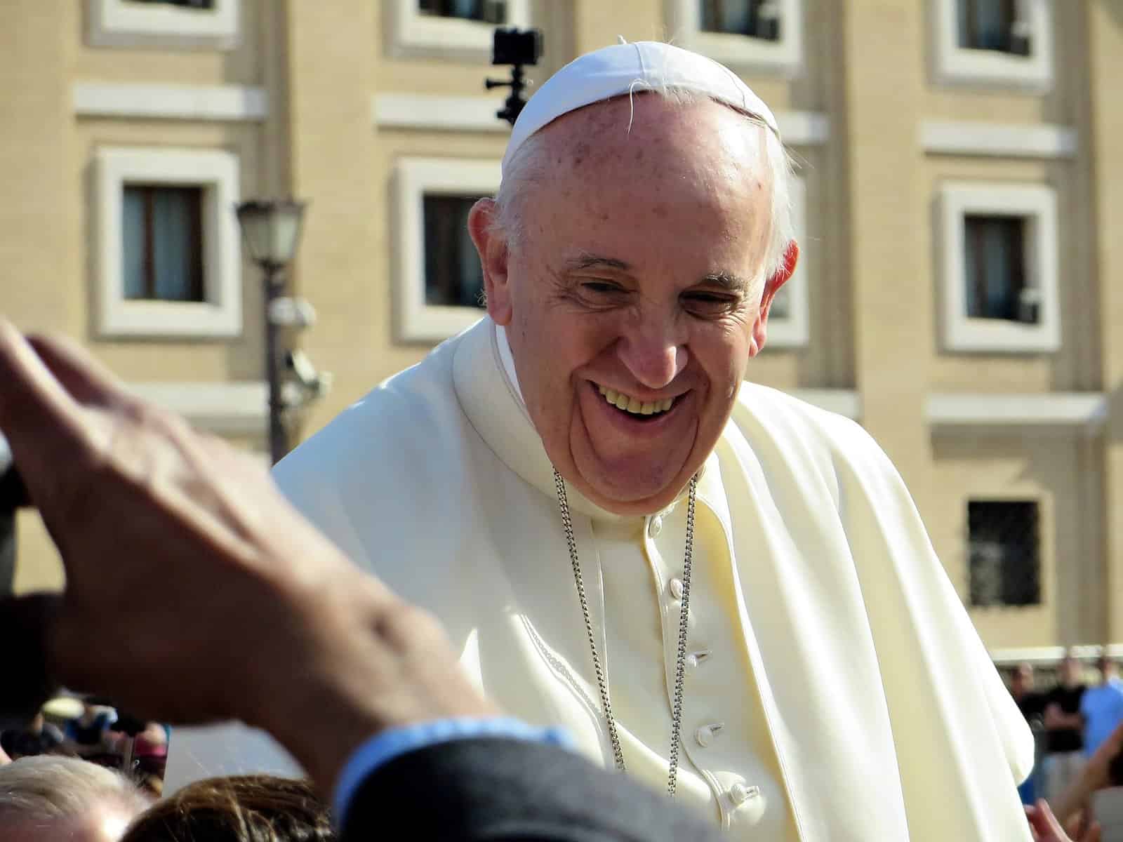 Pope Francis is shown outside facing a crowd and smiling.