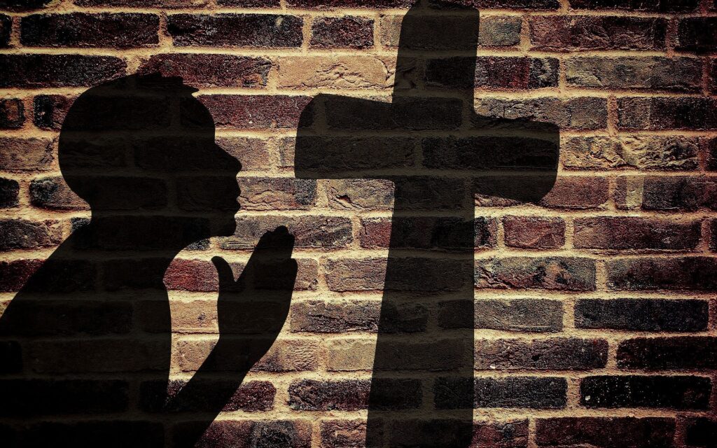 A brick wall has the shadow of a cross on it as the shadown of a man praying with praying hands in front of him.