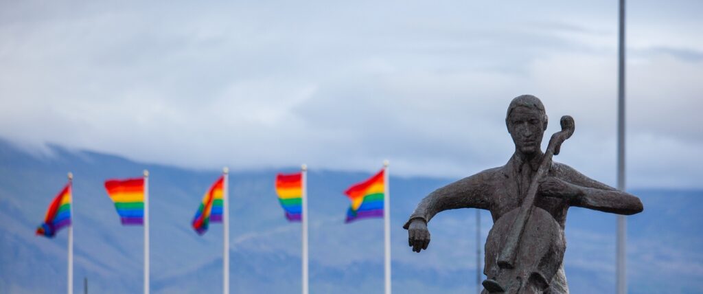 Five rainbow flags are in the distance behind a bronze of a man playing a violin.