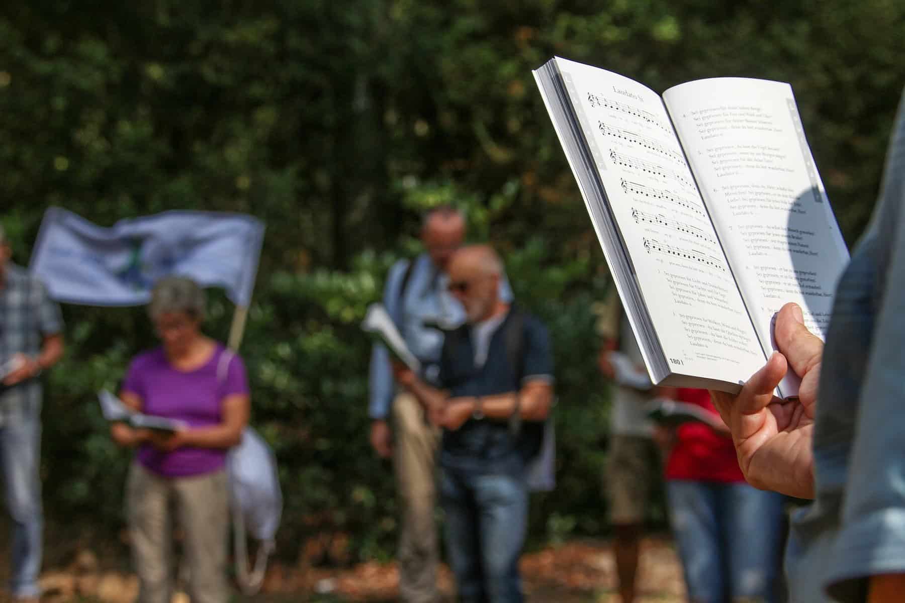 A person holds a songbook in front of them at an outdoor gathering along with others.
