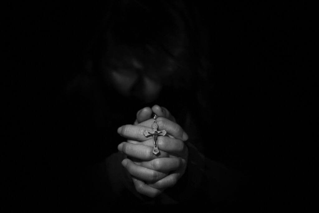 Black and white Image shows only a persons hands clasped together with a crucifix over the knuckles and in a dark room.