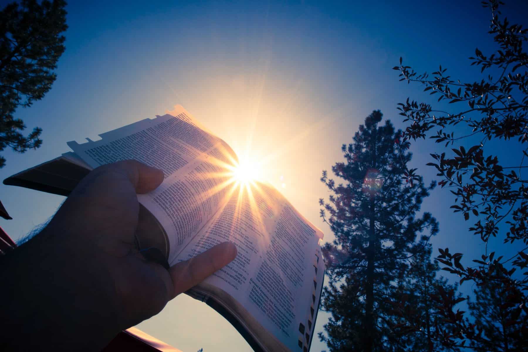 A man's hand holds an open Bible up to the sun and the sun flares go past the page edges.