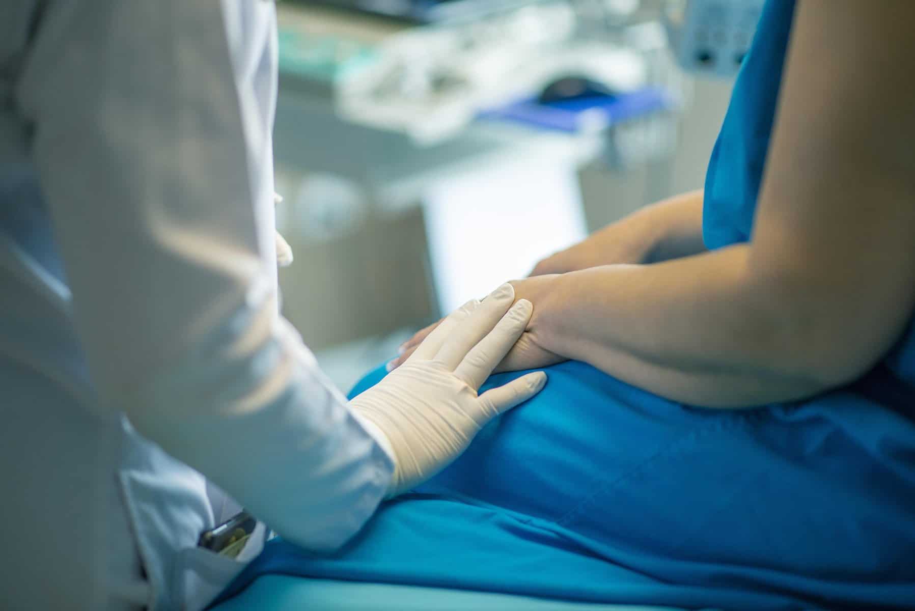 A woman sits on an exam table in a blue gown as the healthcare provider rests a hand on her arm.