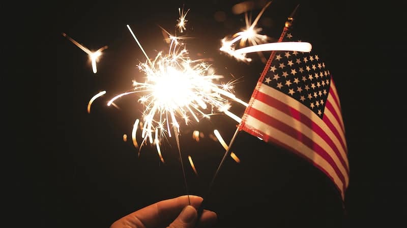 A hand is holding a small American flag against a dark sky with a sparkler behind it.