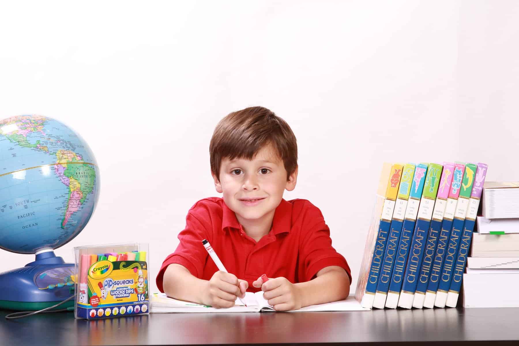A child sits at a desk holding a pen over paper and surrounded by a globe, books, and school supplies.