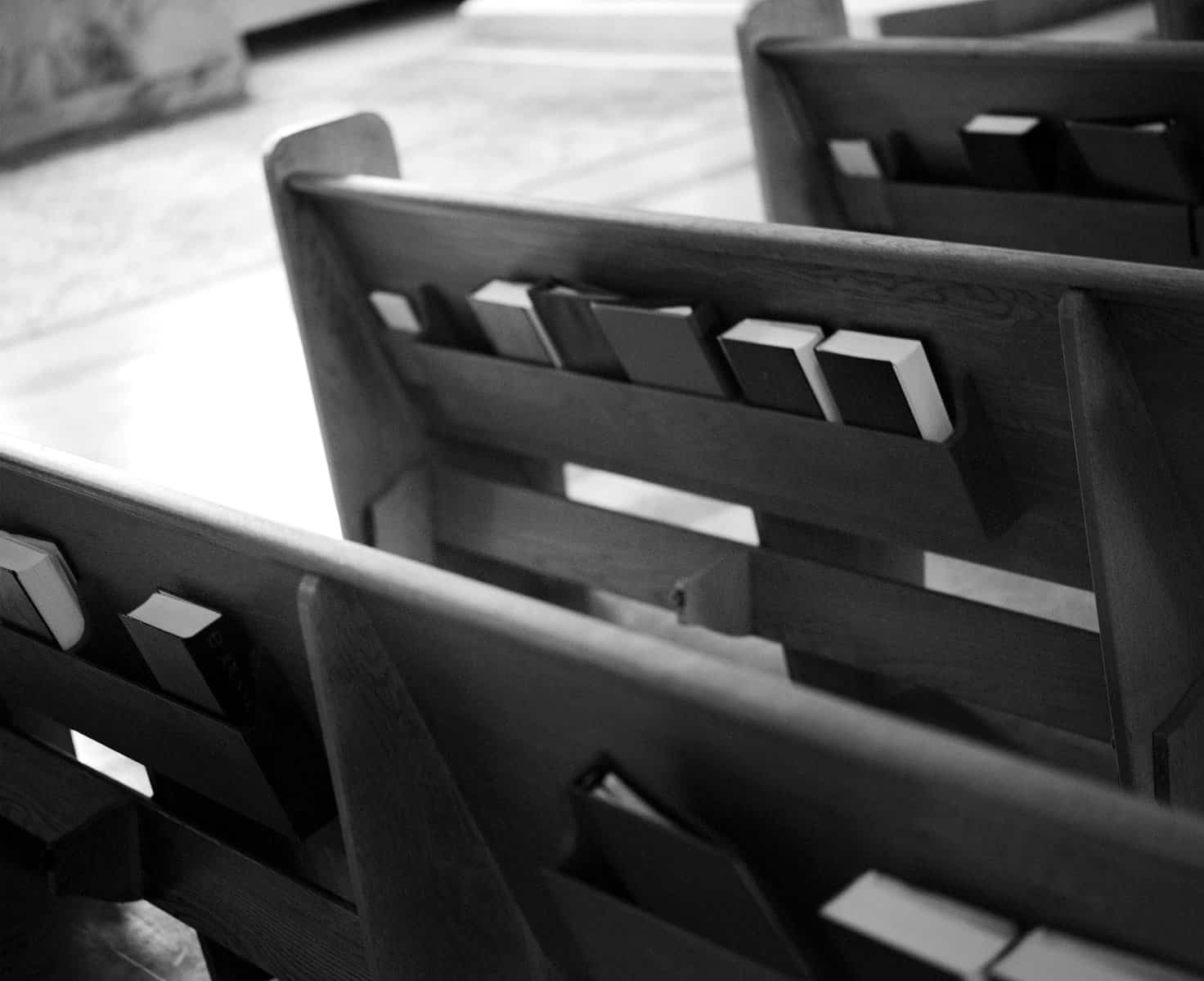 This black and white image shows church pews from the rear side with hymnals and bibles tucked in the back.