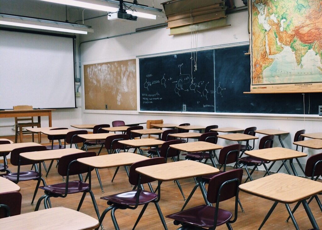 A classroom with a blackboard, large map, and individual desks is shown.
