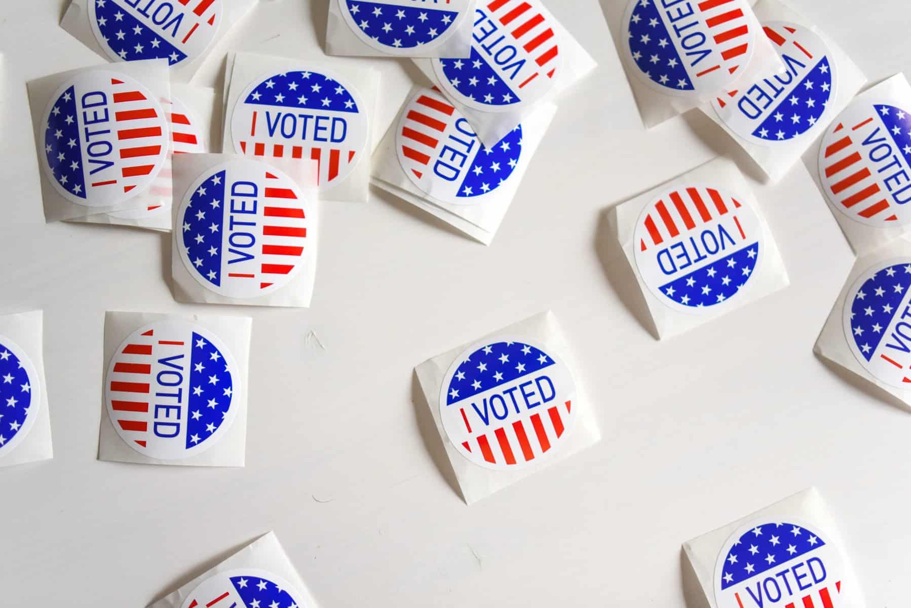 Round red, white, and blue "I voted" stickers are scattered on a white table in soft light.