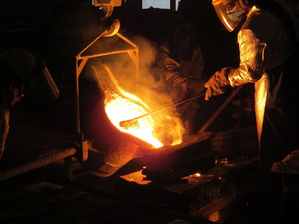 A metal worker wearing protective heavy clothing holds an iron in molten metal.