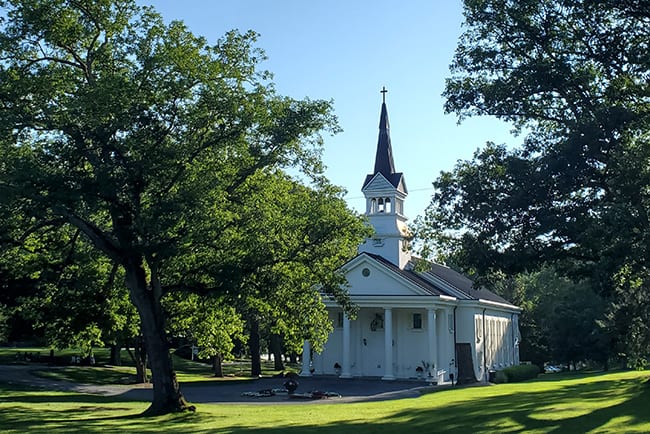A white church with a steeple and surrounded by trees is seen across a green lawn.