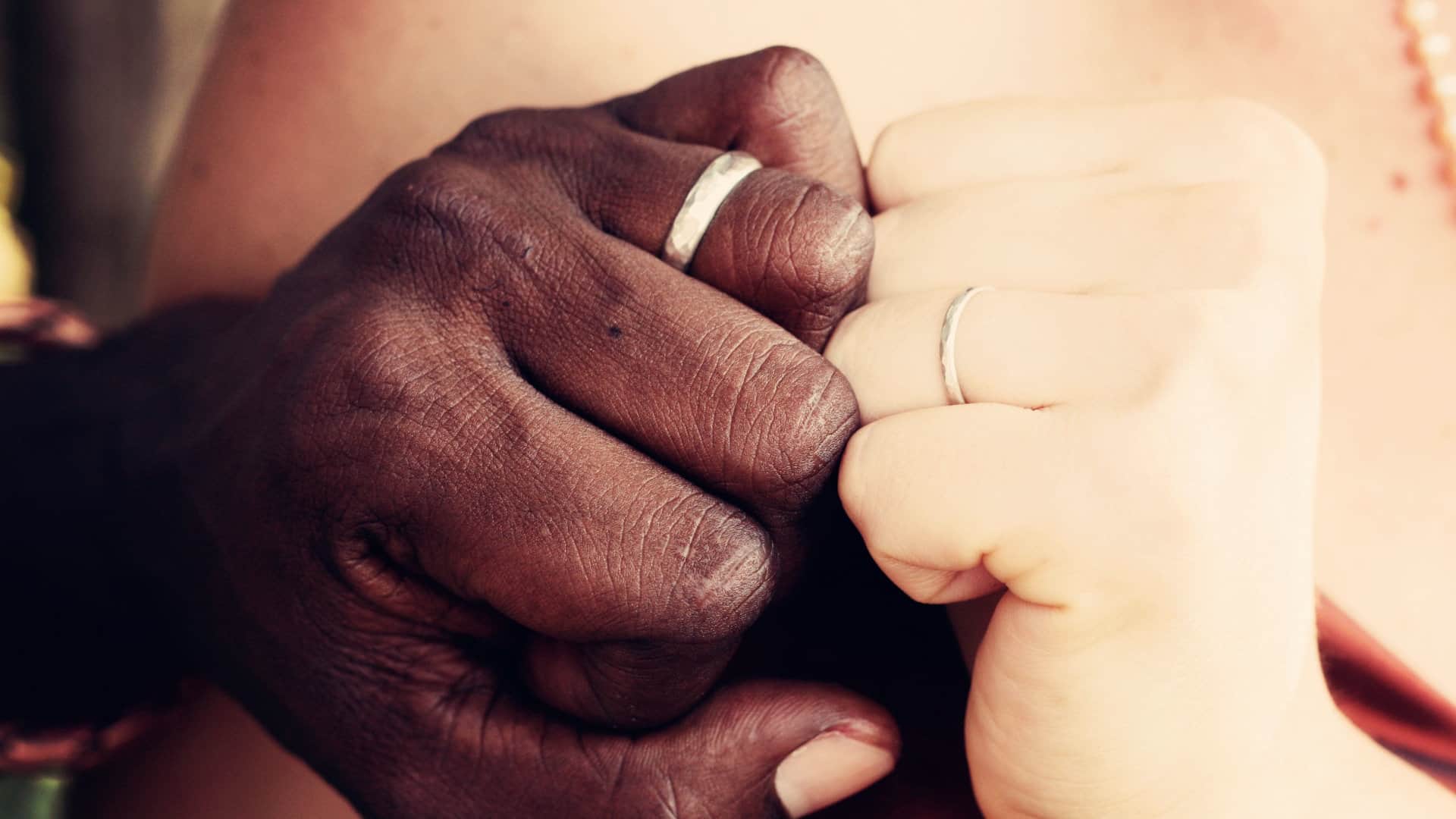 Two hands, one Caucasian, the other Black and wearing wedding rings, are shown up close with knuckles together.