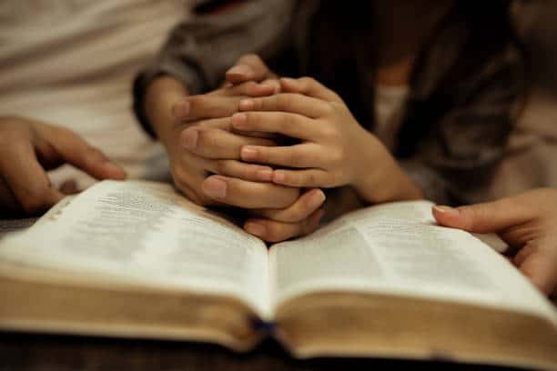 Small hands are clasped and rest on an open bible while adult hands hold the bible's sides.