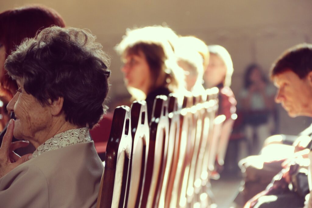 Older people are shown from the side rear seated in rows in a church like setting with the sun coming from behind them.
