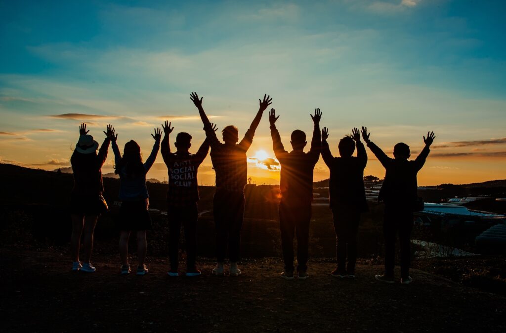 A group of people stand in silhouette with arms raised with the sunset behind them.