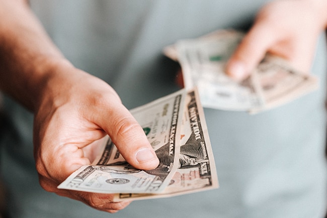 A man's hands are shown holding paper money in each hand with one extended as if handing the money to someone.