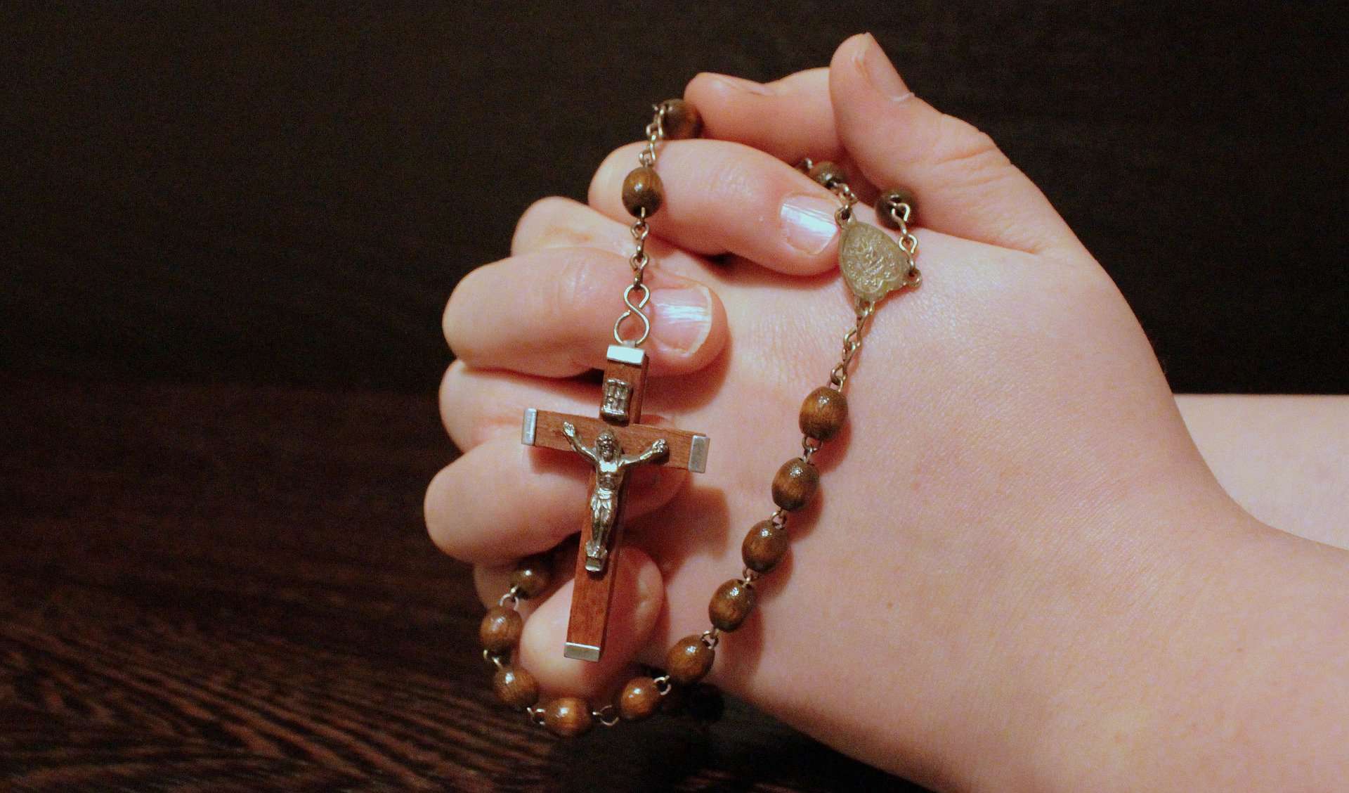A child's clasped hands are seen from the side with a rosary and cross draped over them.
