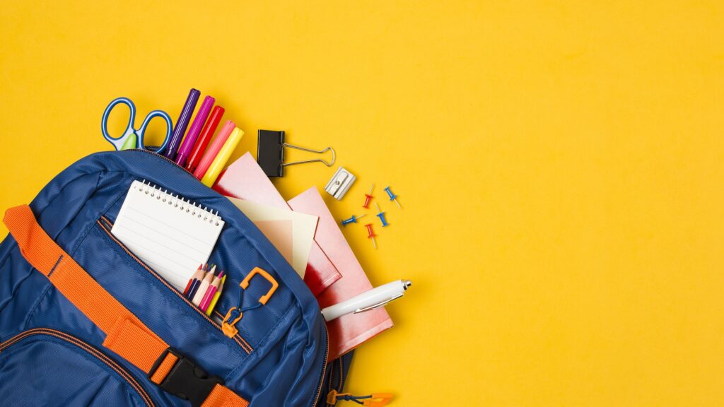 A blue backpack lays on a bright yellow background with school supplies spilling out of it.