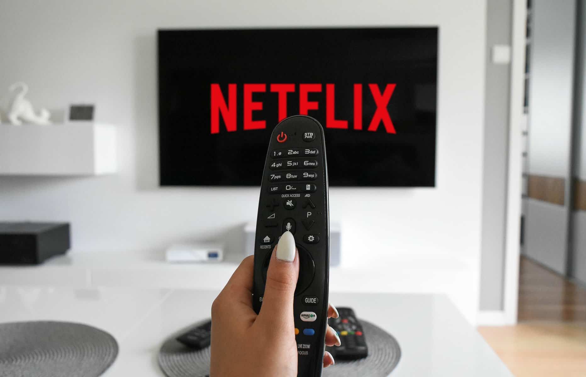 A TV with the Netflix logo hangs on a white wall with a woman's manicured hand holding a remote between the camera and the tv.
