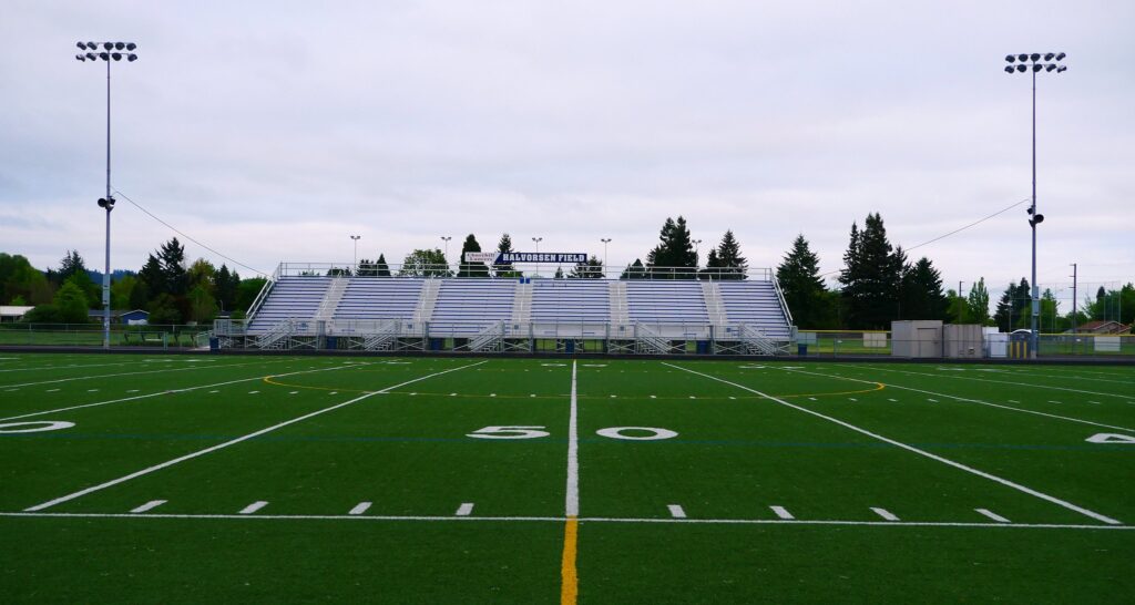 A football field is shown in low light from the sidelines with bleachers across the field.