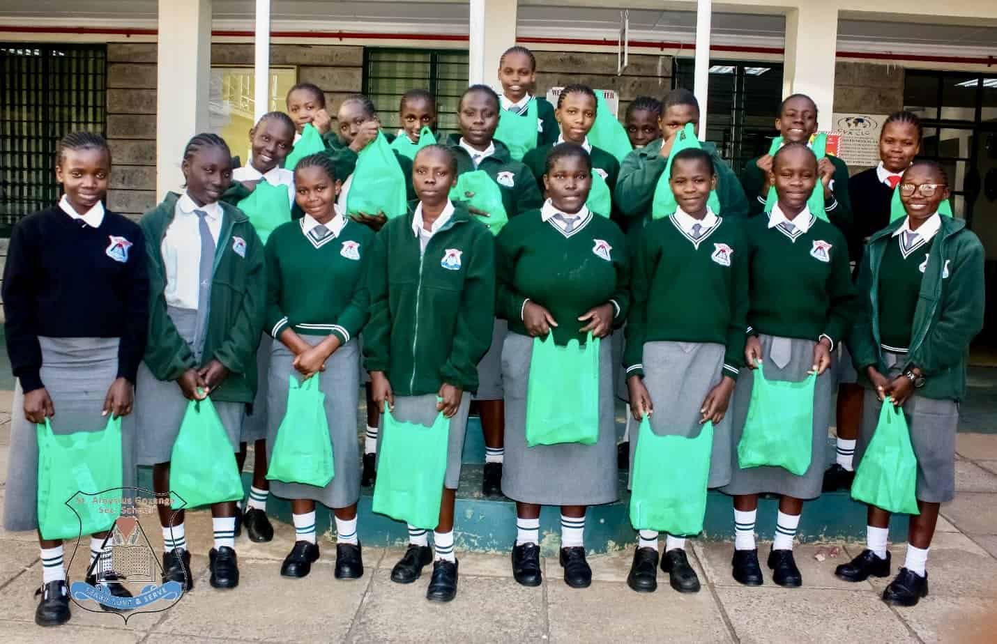 African girls stand in a group dressed in school uniforms and holding green plastic bags.