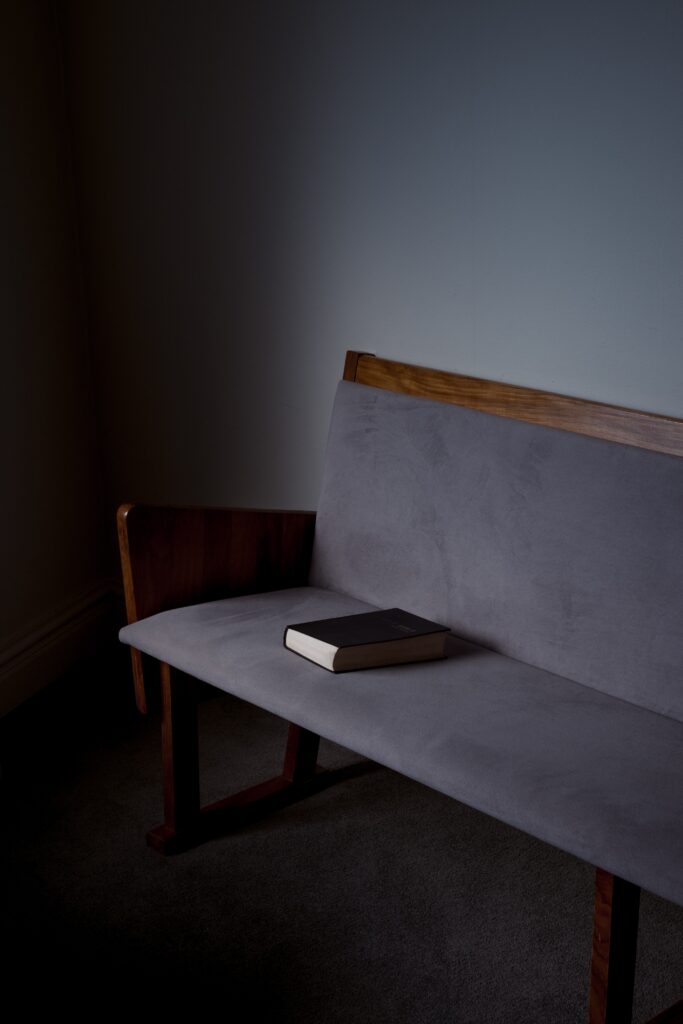 A black covered hymnal or Bible is shown at the end of a dark grey fabric covered pew with very soft natural light illuminating it.