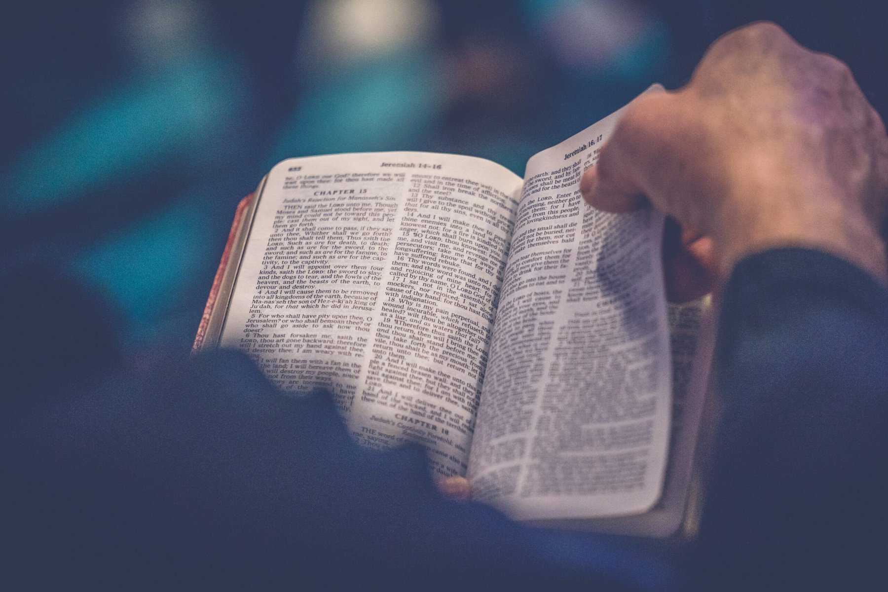 An open Bible is shown close up sitting on a man's lap with his right hand ready to turn the page.