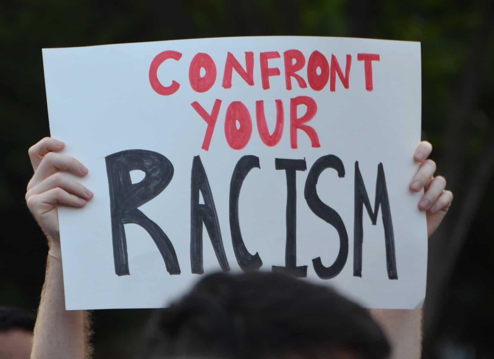 A person's hands are shown holding a white sign up with the words "Confront your racism" written on it.