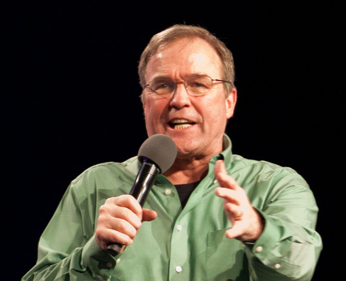 Mike Bickle is seen in a green shirt and speaking into a microphone he's holding.