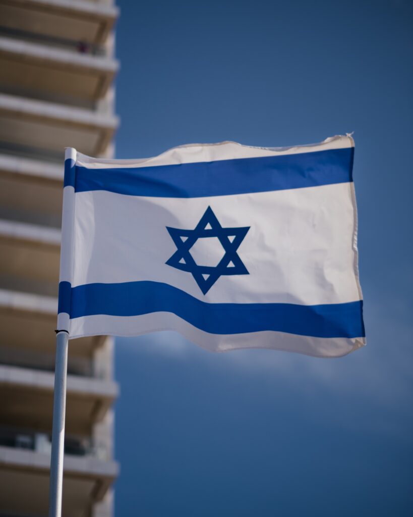 Israel's flag is shown waving in the wind with the corner of a high-rise building as a backdrop.