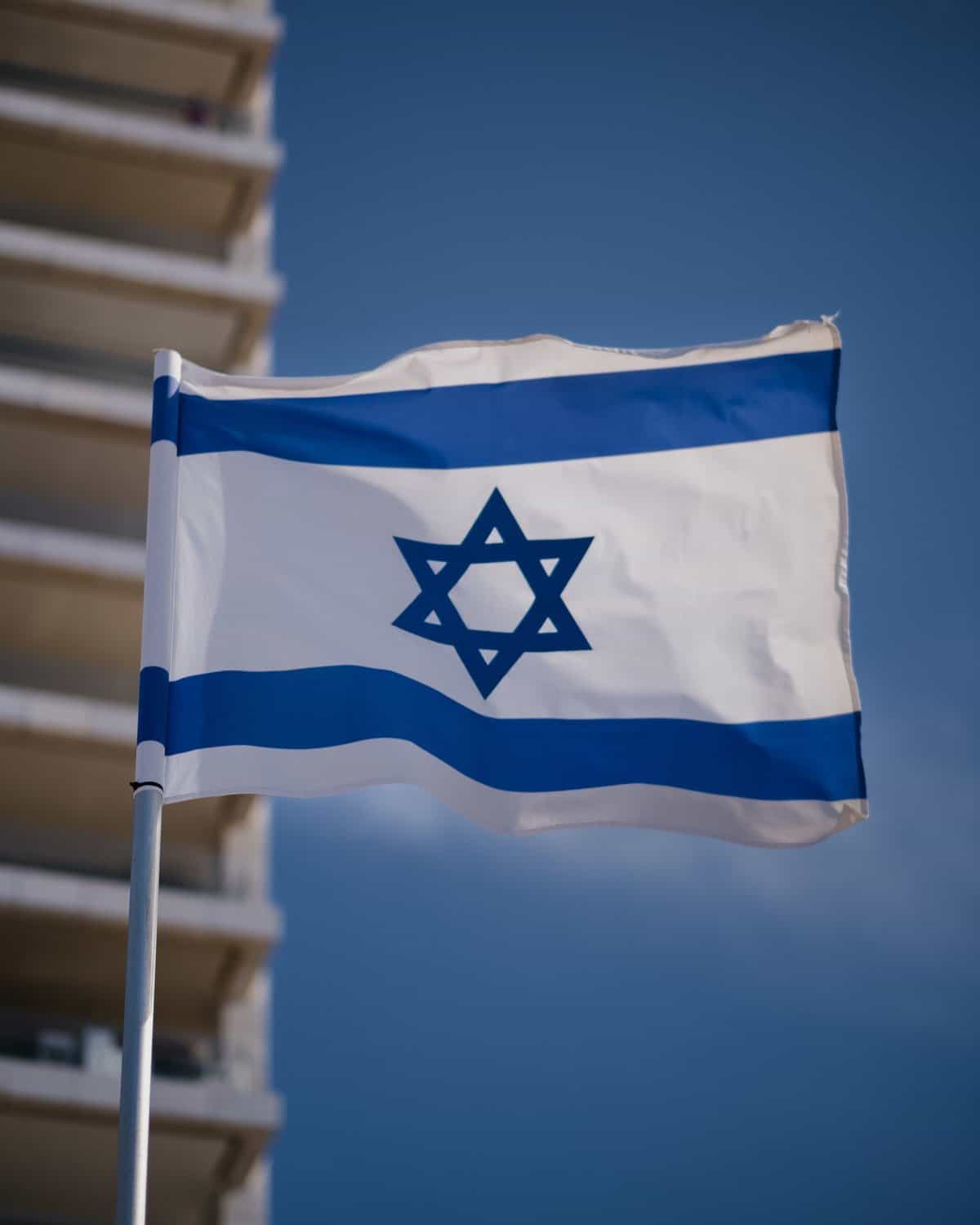 Israel's flag is shown waving in the wind with the corner of a high-rise building as a backdrop.