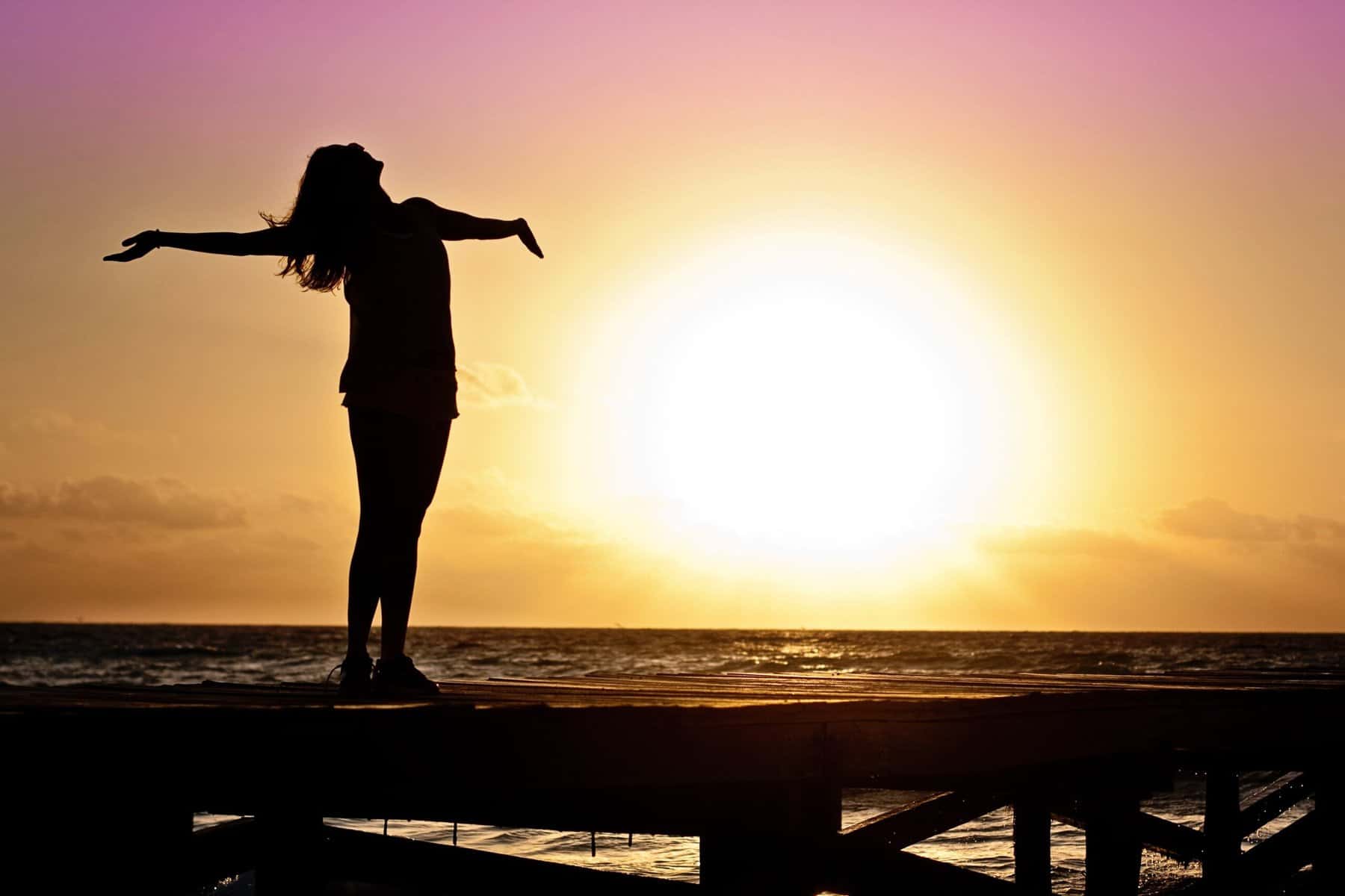 A woman seen in silhouette stands facing the sun and ocean at sunset with her arms spread and head tilted back.