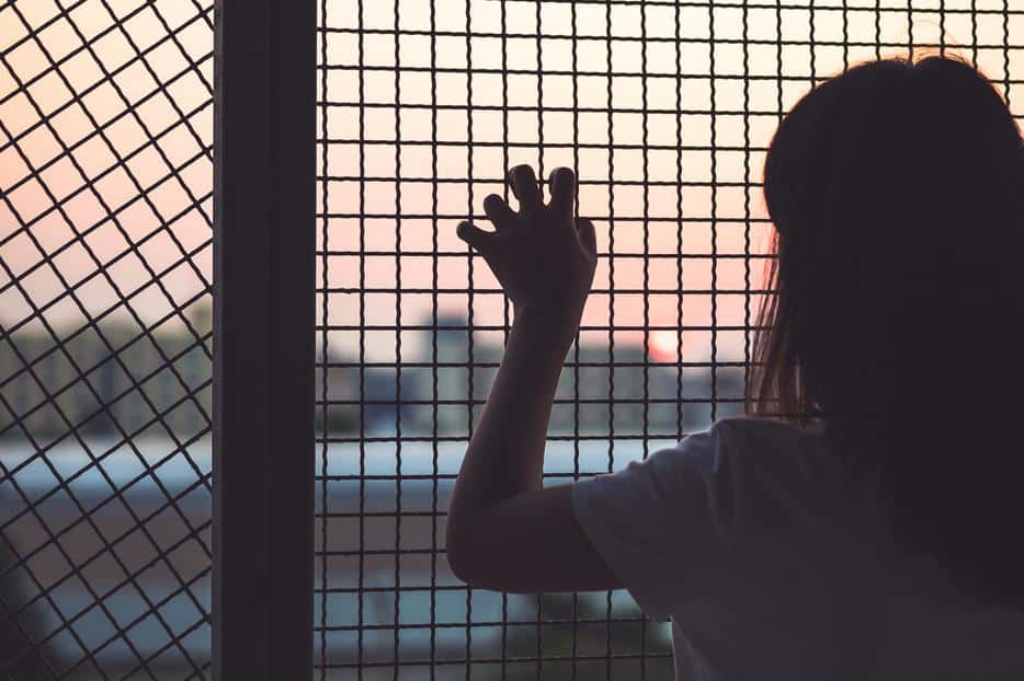 A young girl stands in silhouette with back to camera and a hand gripping a metal wire fence.