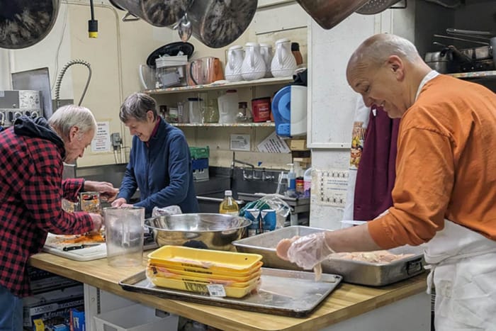 Four older people are seen preparing a meal in a community type kitchen.