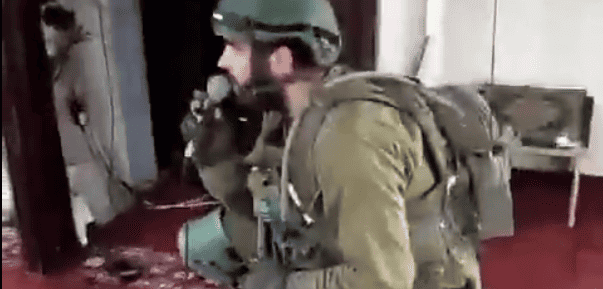 A soldier in full gear crouches and speaks into a microphone.