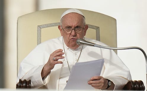 Pope Francis sits in a papal chair and speaks into a microphone while holding some paper.